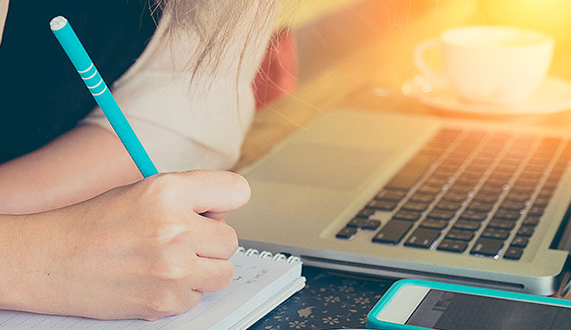 Fotografia de persona tomando escribiendo en un cuaderno, con computador y teléfono sobre la mesa