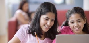 Imagen con dos niñas trabajando en un computador. Una está utilizando el computador y la otra toma apuntes en un cuaderno