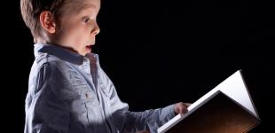 La imagen muestra a un niño en fondo negro mirando un libro abierto entre sus manos con cara de asombro