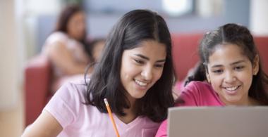 Imagen con dos niñas trabajando en un computador. Una está utilizando el computador y la otra toma apuntes en un cuaderno