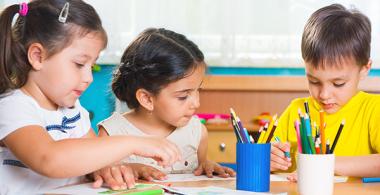 La imagen muestra a dos niñas y un niño realizando una actividad en una mesa escolar